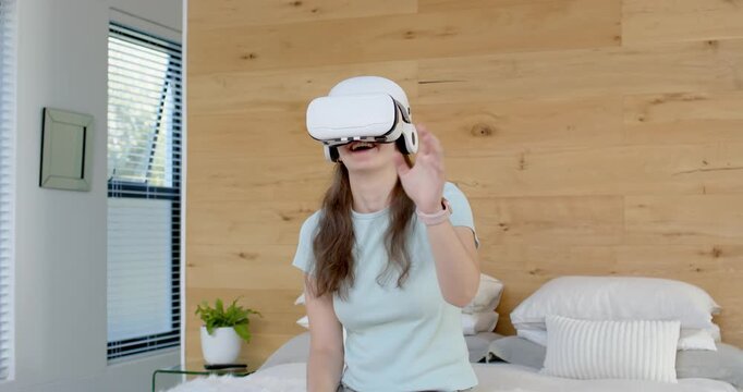 Adult woman sitting in bedroom on bed, wearing white VR-headset and smartwatch, navigating VR menu
