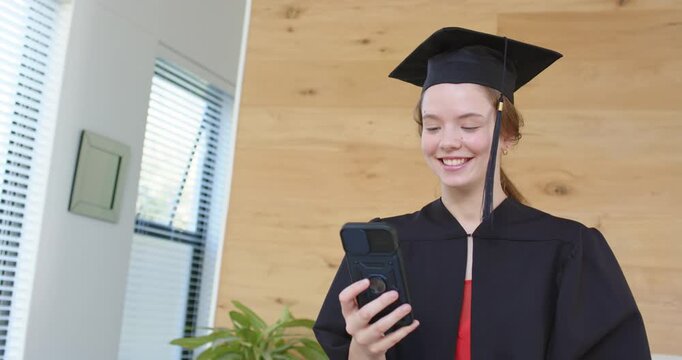 Gowned adult female getting call marking grad holding phone at home waving adjusting cap tassel