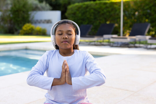 African American youth female sitting meditating on pool deck wearing white over-ear headphones