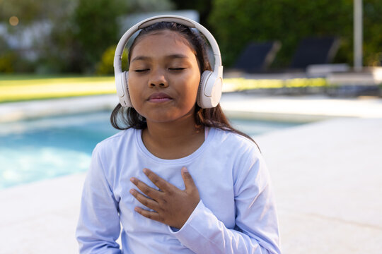 Female child sitting at pool edge on pool deck wearing white headphones and light long-sleeve shirt