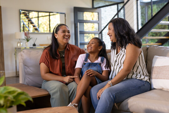 Diverse female family with child laughing while sitting on beige sofa by coffee table and plant