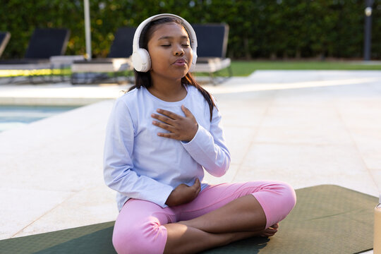 African American child sitting on mat by pool wearing blue top, pink pants, headphones, bottle near