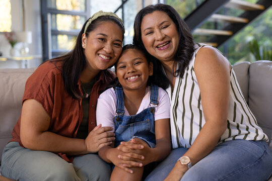 Female family of three sitting on living room sofa, smiling and leaning toward child in overalls