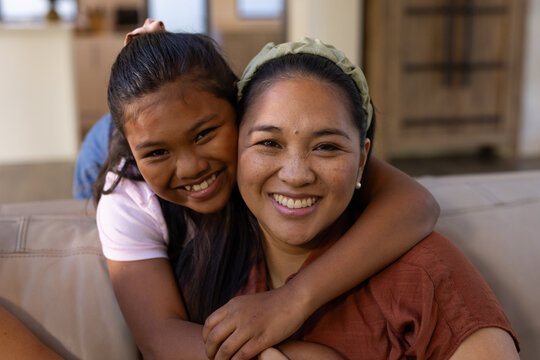 Diverse mother hugging teenage daughter on light-colored sofa at home, wearing rust top