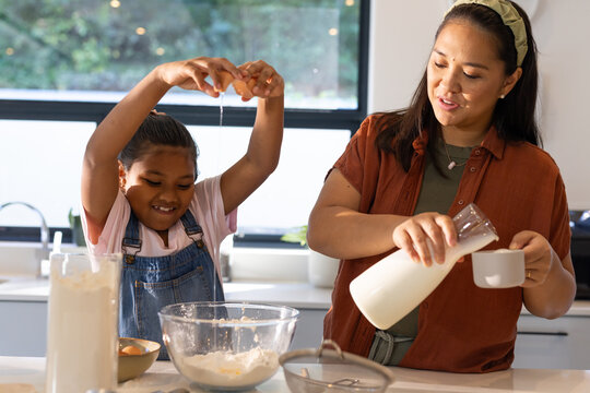Diverse female mother and child, child in overalls cracking egg and pouring milk in kitchen bowl