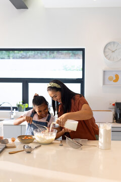 Diverse mother and youth daughter pouring milk and stirring batter in home kitchen with mixing bowl