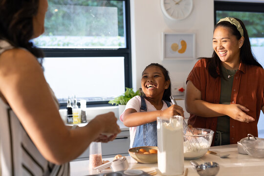 Diverse female family helping child in overalls mixing batter on kitchen island with glass bowl