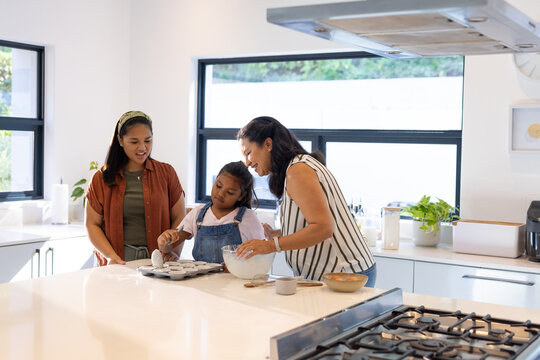 Diverse female family baking muffins on white countertop island in modern kitchen using glass bowl