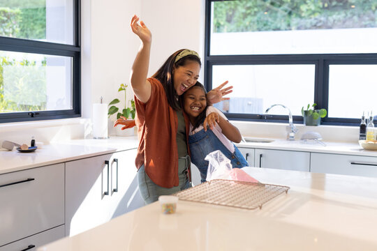 Diverse mother and youth daughter hugging, baking at island with sprinkles, daughter in overalls