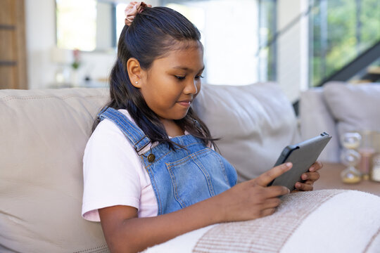 African American child sitting on beige couch at home in denim overalls holding dark tablet
