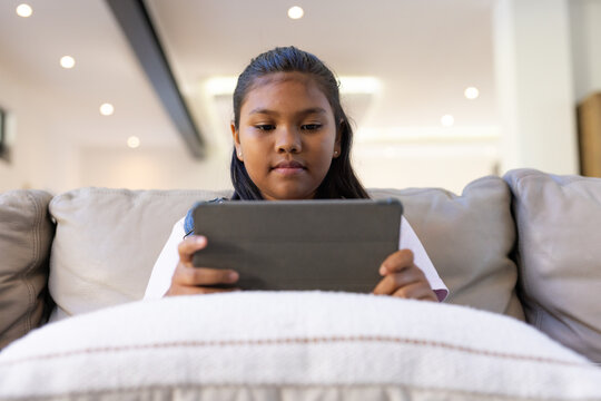 Adolescent African American girl sitting on beige sofa at home wearing white shirt holding tablet
