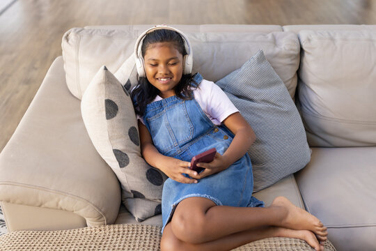 Female child reclining on beige sofa at home, wearing denim overall and headphones, holding phone
