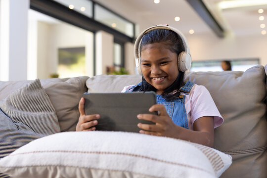 African female child sitting on sofa wearing white headphones and denim overalls, holding tablet