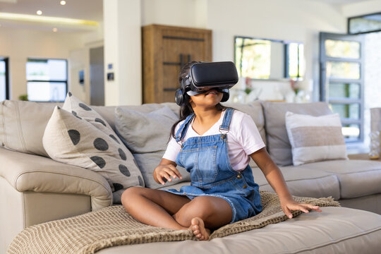African American school-age girl sitting on sofa at home, wearing VR headset and denim overalls