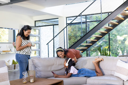 Diverse female family lying, leaning and standing in modern living room with smartphone and mug