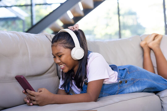 African American pre-teen girl lying on light gray couch wearing headphones, holding red-pink phone
