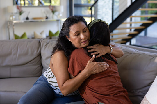 African American mother and Asian daughter hugging on light gray leather sofa, wristwatch visible
