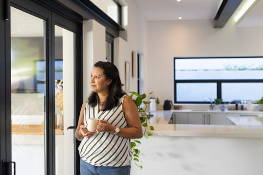 Mature African American woman in striped top holding mug gazing through black doors in kitchen