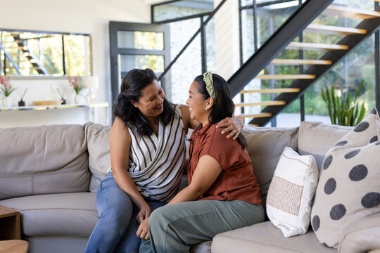Diverse female mother and daughter sitting on beige sectional sofa in living room, smiling