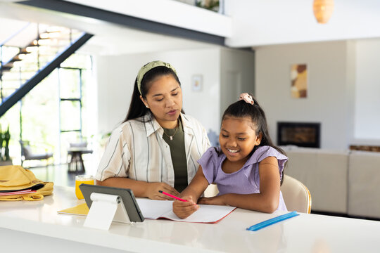 Asian mom guiding school-age African American daughter doing homework at kitchen island with tablet