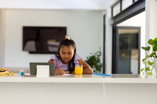 African American school-age girl sitting at kitchen island, writing with pink pencil, using tablet