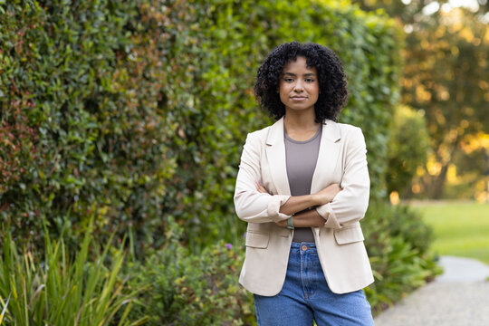 Woman standing with arms crossed on gravel walkway near manicured hedge wearing blazer and bracelet