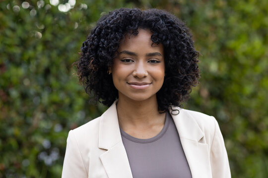 Woman standing facing camera in garden wearing beige blazer, taupe-gray top, hoop earrings