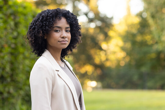 Woman standing facing camera in garden wearing beige blazer and taupe top, hedge and lawn behind