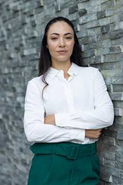 Woman leaning against textured stacked stone wall, wearing white blouse and green belted trousers
