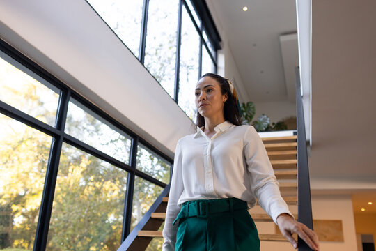 Woman walking down staircase at home by windows wearing white blouse and teal pants, copy space