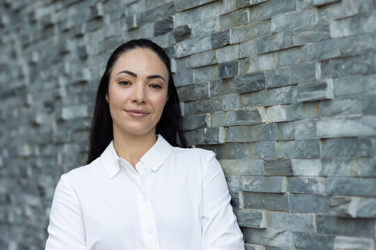 Adult woman leaning on stacked stone wall, wearing white button-up shirt, crossing arms, copy space
