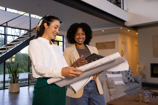 Diverse female colleagues reviewing rolled architectural plans and using tablet in living room