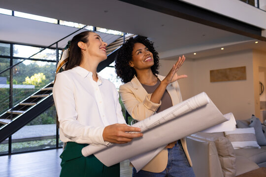Two women in business-casual clothes holding rolled plans and pointing in living room near stairs