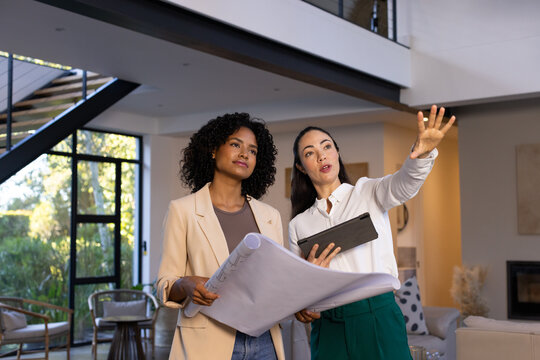 Female colleagues standing in modern loft, holding tablet and plans, wearing blazer and blouse