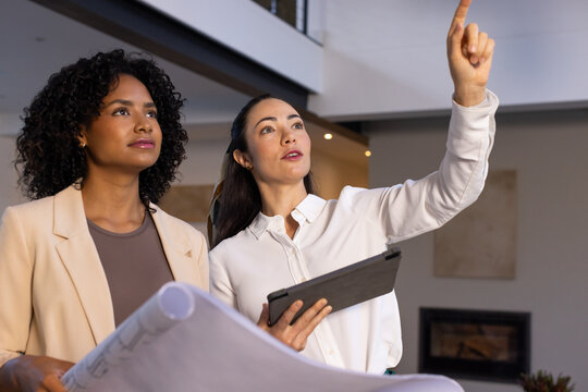 Diverse female coworkers holding blueprints and tablet, pointing and inspecting modern mezzanine