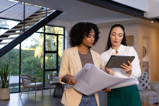Diverse female colleagues reviewing rolled plan and tablet in business attire in modern home