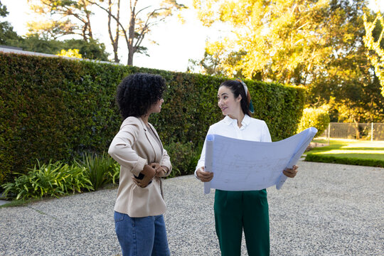 Diverse female colleagues in blazers reviewing design plans on driveway by hedge, smartphone at hip