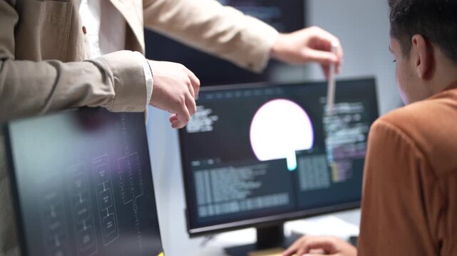 A senior technology architect pointing out system optimization parameters and database structure models on a monitor to a software developer.