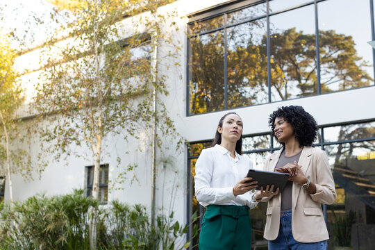 Diverse female coworkers talking over tablet outside office, wearing blouse and watch, copy space