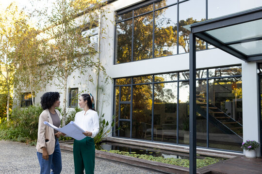 Diverse female colleagues reviewing blueprints at two-story house with water trough, copy space