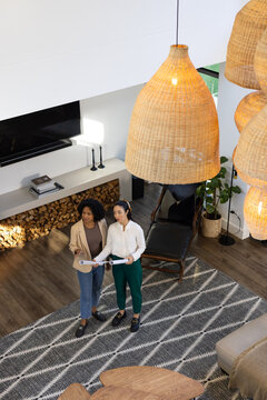 Diverse female coworkers reviewing rolled plans in modern living room with woven pendant lights