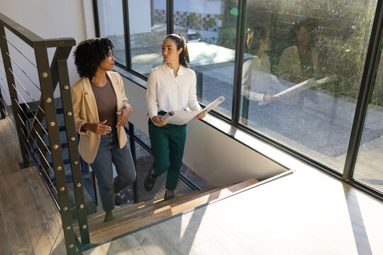 Diverse female coworkers walking up wood stairs by window with rolled plans checking cable railing