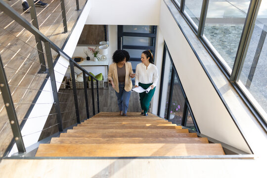 Two adult women walking up wooden stairs in loft, wearing blazers and holding rolled documents