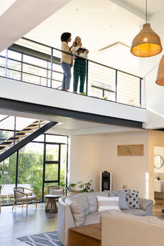 Diverse female coworkers standing on mezzanine balcony overlooking living room, holding tablet