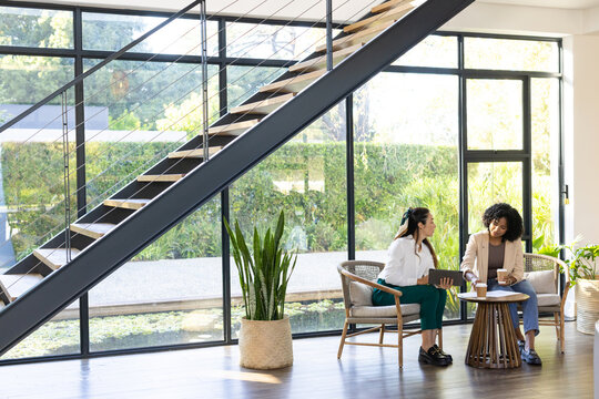 Diverse female coworkers sitting in modern coworking lounge reviewing tablet and papers, copy space