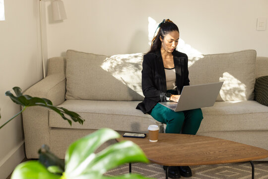 Woman sitting on sofa typing on laptop at home with plants, phone, cup, wearing black blazer