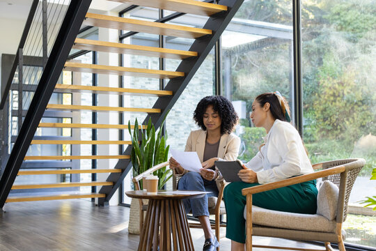 African American and Asian women in blazers reviewing printed documents and tablet in office lounge