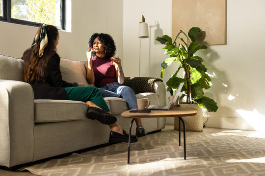 Diverse female friends talking on sofa in living room with laptop, mug, burgundy top, blazer