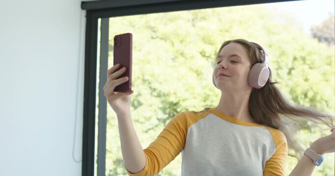 Woman holding smartphone out, flipping hair, adjusting pink headphones, filming near large window