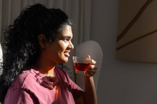 Indian woman sitting in living room holding clear glass cup of tea, maroon T-shirt, copy space